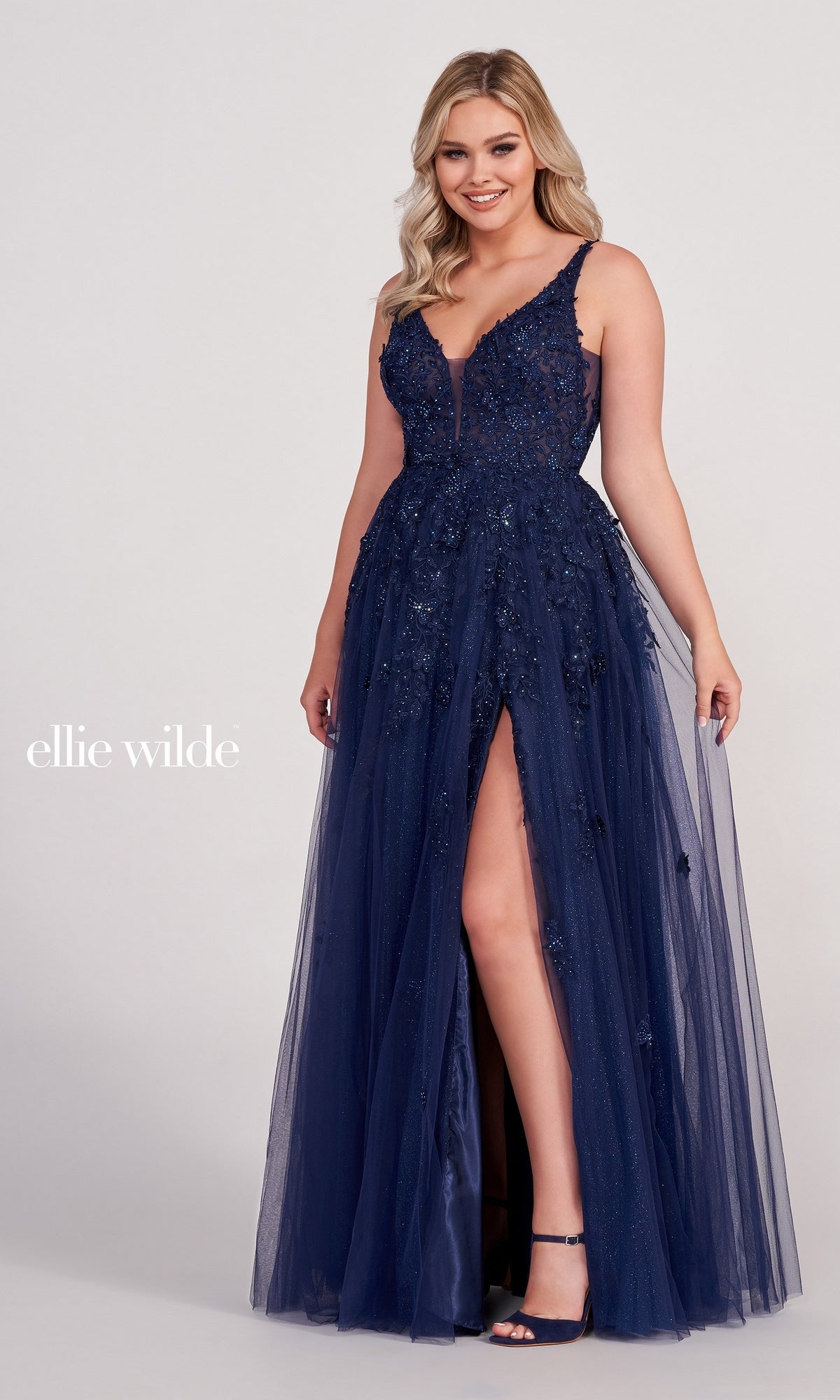 A woman wears the Ellie Wilde Side-Slit Long Prom Ball Gown EW34103 in navy blue, featuring floral embellishments, a sheer neckline, sleeveless design, and a thigh-high slit for an elegant evening look.